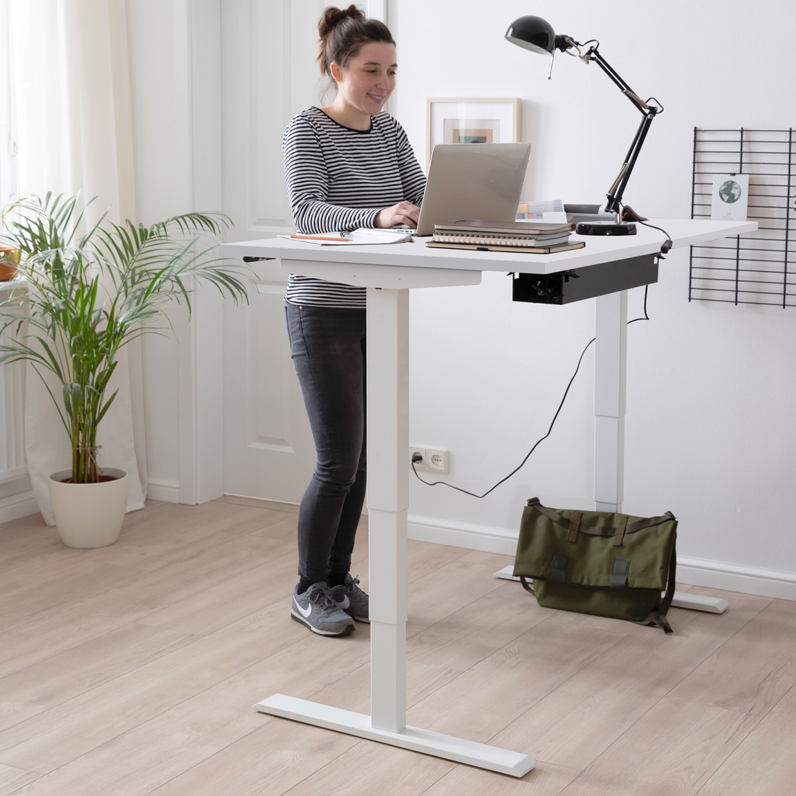 Frau arbeitet stehend an einem weißen höhenverstellbaren Schreibtisch mit Laptop, Schreibtischlampe und Notizbüchern in einem hell eingerichteten Homeoffice.