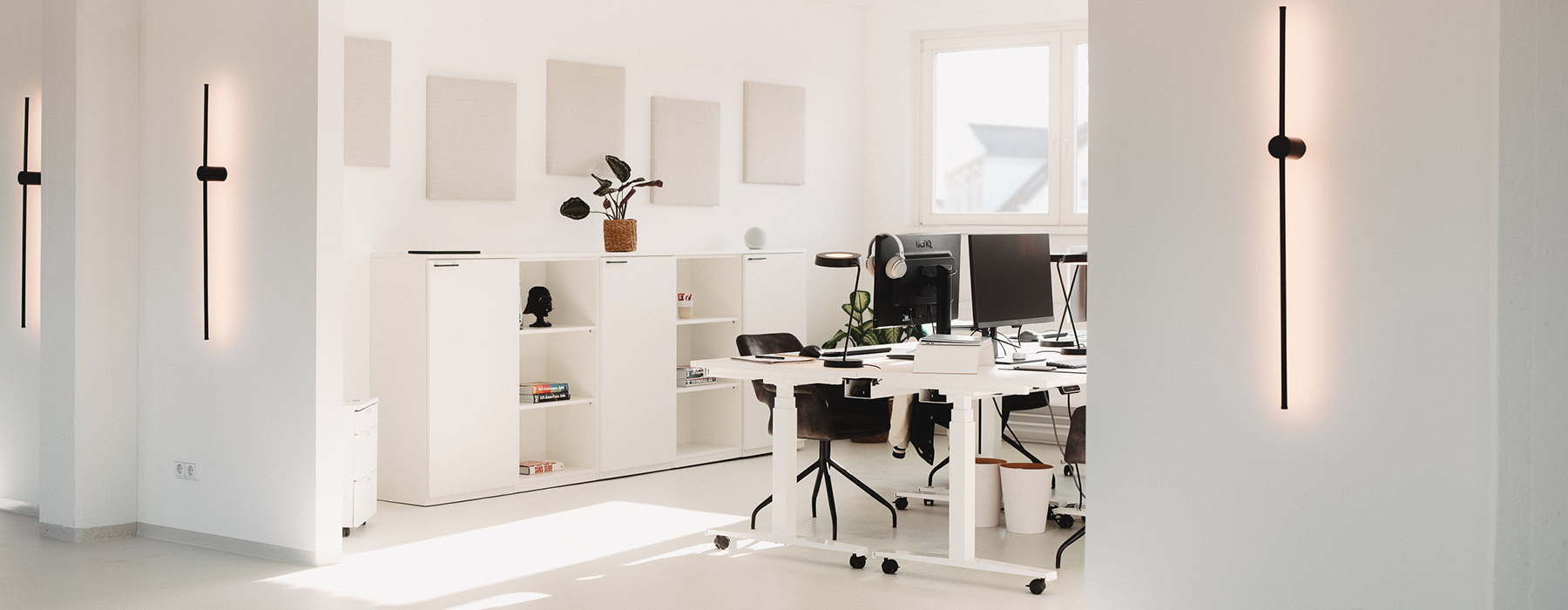 Modernes Büro mit weißen höhenverstellbaren Schreibtischen, ergonomischen Stühlen und minimalistischer Dekoration, beleuchtet von stilvollen Wandlampen.